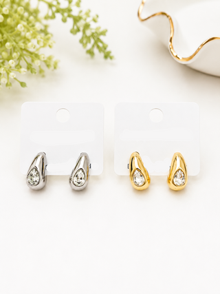 Two pairs of teardrop-shaped stud earrings on display cards. One pair is silver-toned and the other is gold-toned, both featuring a clear teardrop gemstone.