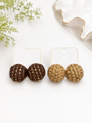Two pairs of woven round stud earrings on display cards. One pair is dark brown and the other is light tan, both with gold-toned metal edges and displayed on white cards with the brand name 'ITS' in gold.